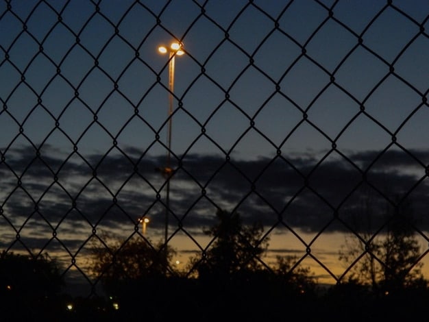 A photo showing a close-up of the US-Mexico border fence at night. The image has a strong focus on the texture of the fence, with spotlights illuminating the structure and hinting at a high-security environment.