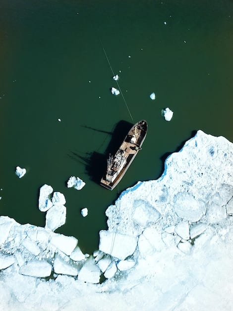 An aerial shot of a large cargo ship navigating through icy waters in the Arctic. The ship is surrounded by chunks of floating ice, and the landscape shows a mix of snow-covered mountains and icy plains.